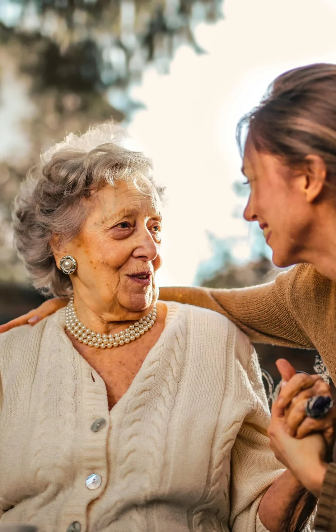 An older woman in a cream cardigan smiling at a younger woman beside her